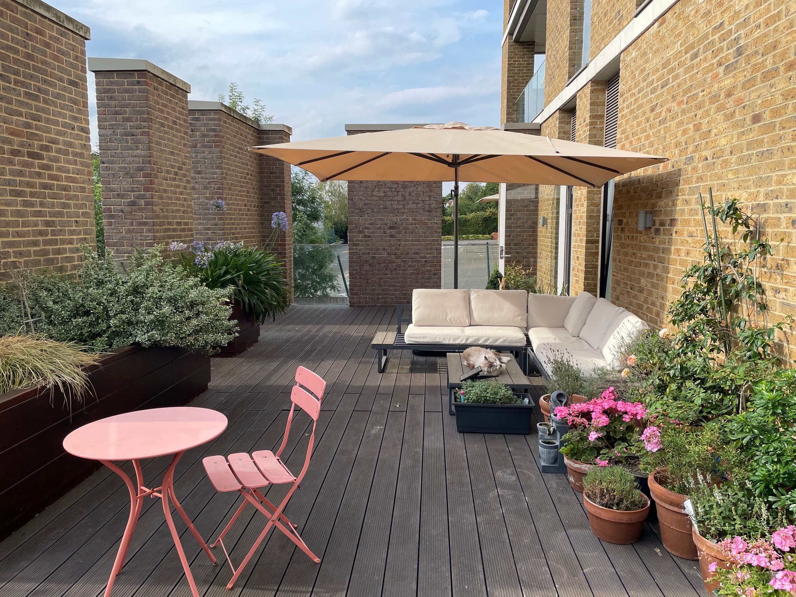 The terrace in afternoon sun with a corner sofa beneath a parasol, a bistro table and planting along the wall